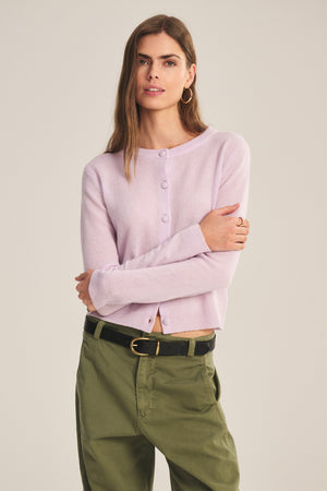 A person stands with arms crossed against a plain background, wearing the NOLIE CASHMERE CARDIGAN by Velvet by Graham & Spencer in light purple and olive green pants.
