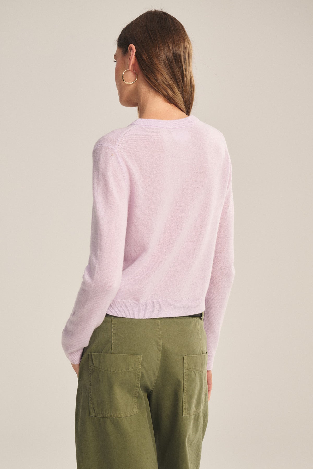 A person with long brown hair, facing away, wears the NOLIE CASHMERE CARDIGAN by Velvet by Graham & Spencer in light pink, paired with green pants featuring large back pockets.