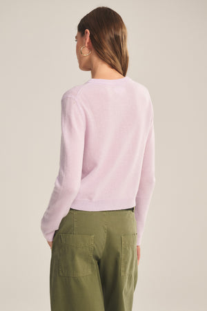 A person with long brown hair, facing away, wears the NOLIE CASHMERE CARDIGAN by Velvet by Graham & Spencer in light pink, paired with green pants featuring large back pockets.