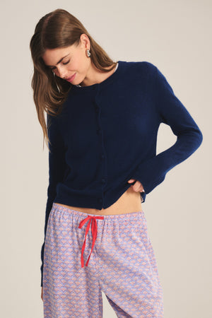 A woman wears the Velvet by Graham & Spencer NOLIE CASHMERE CARDIGAN in navy blue with patterned pajama pants featuring a red drawstring, standing against a plain background.