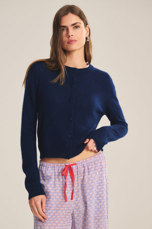 A woman wears the NOLIE CASHMERE CARDIGAN by Velvet by Graham & Spencer in navy blue, paired with pink patterned pajama pants with a red drawstring, standing against a neutral background.