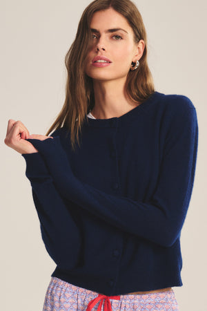 A woman wearing the NOLIE CASHMERE CARDIGAN by Velvet by Graham & Spencer in navy blue, paired with patterned pants, stands against a plain background.