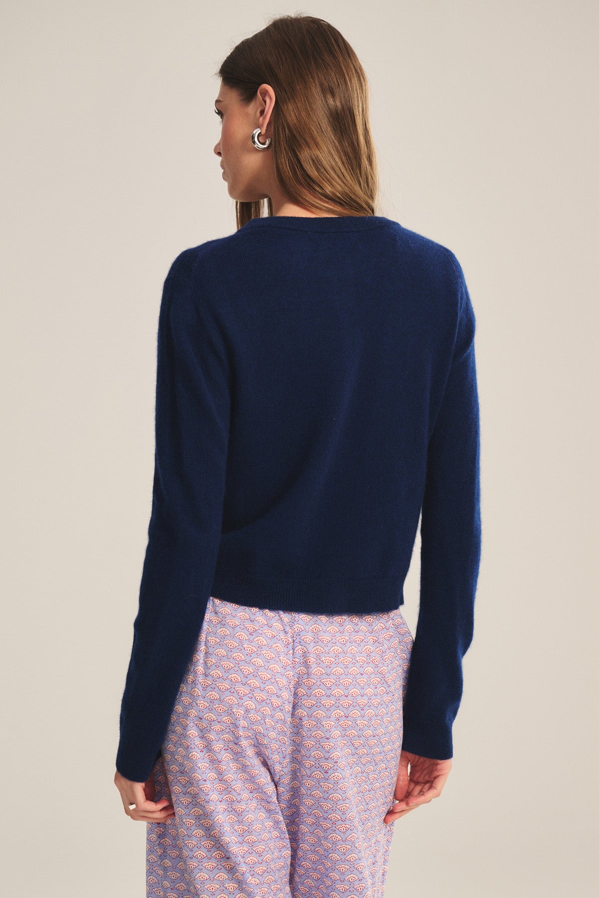 A person stands facing away, wearing the NOLIE CASHMERE CARDIGAN by Velvet by Graham & Spencer in navy blue and light pink patterned pants.-40808678817985