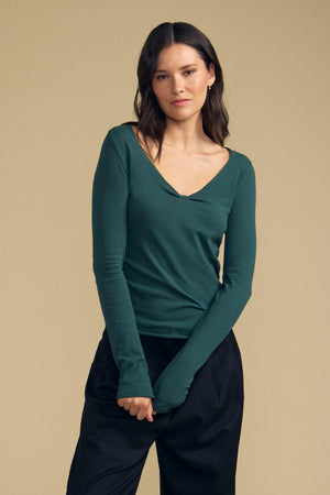A woman stands against a beige background, wearing the NOURA TEE by Velvet by Graham & Spencer—a dark green, long sleeve cotton jersey V-neck paired with black pants—her arms relaxed in front of her.