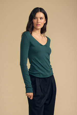 A woman stands against a beige background wearing the NOURA TEE by Velvet by Graham & Spencer—a long-sleeve dark green cotton jersey top with a subtle scoop V-neckline—paired with loose black pants, her head turned slightly to the side.