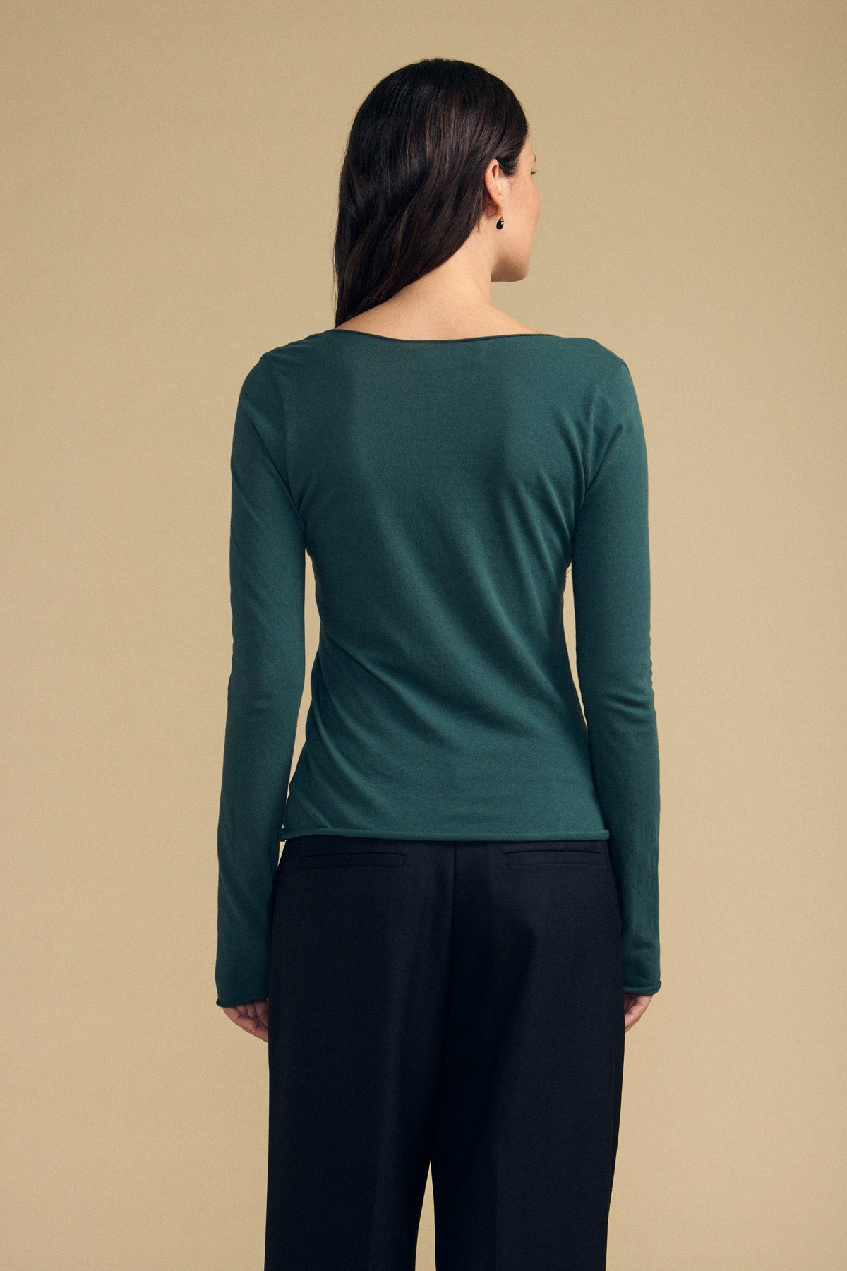 A person with long dark hair, seen from behind, is wearing the Velvet by Graham & Spencer NOURA TEE—a dark green V-neckline long sleeve classic women's shirt—paired with black pants against a beige background.-41838211956929