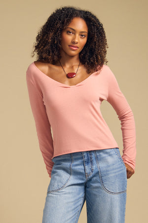 A woman with curly hair stands against a plain beige background wearing the NOURA TEE by Velvet by Graham & Spencer, a pink cotton jersey top featuring a scoop V-neckline and long sleeves, paired with blue jeans and a red pendant necklace.