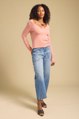 A woman stands in a neutral setting wearing the NOURA TEE by Velvet by Graham & Spencer, a pink cotton jersey long sleeve top with a scoop v-neckline, paired with loose blue jeans, brown heeled sandals, and a red necklace.