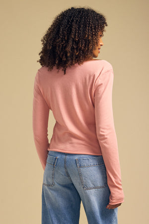 A person with curly hair stands facing away against a neutral background, wearing the NOURA TEE by Velvet by Graham & Spencer—a long sleeve cotton jersey tee featuring a scoop v-neckline in pink—paired with light blue jeans.