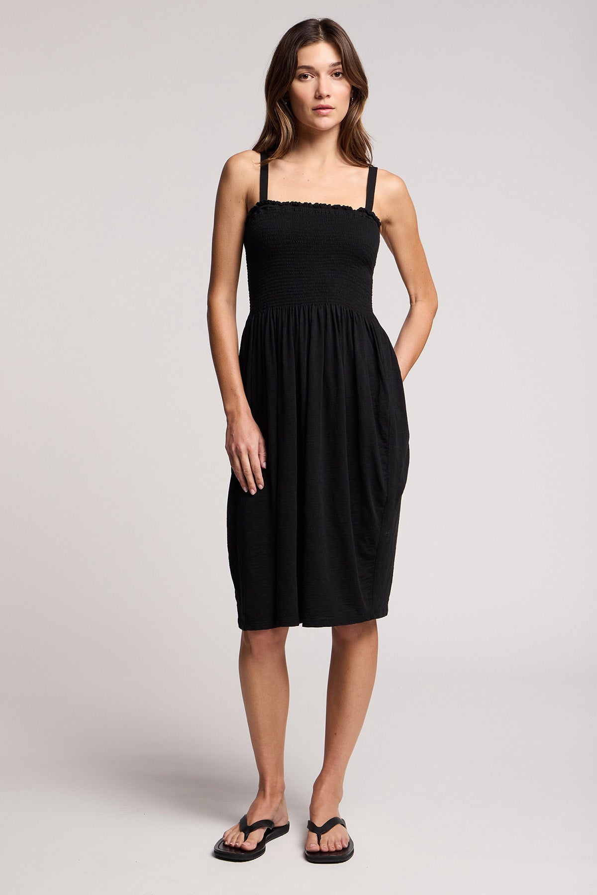 A woman faces forward in the CRESSIDA DRESS by Velvet by Graham & Spencer, a black sleeveless summer dress paired with black flip-flops, set against a plain light background.