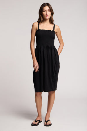 A woman faces forward in the CRESSIDA DRESS by Velvet by Graham & Spencer, a black sleeveless summer dress paired with black flip-flops, set against a plain light background.
