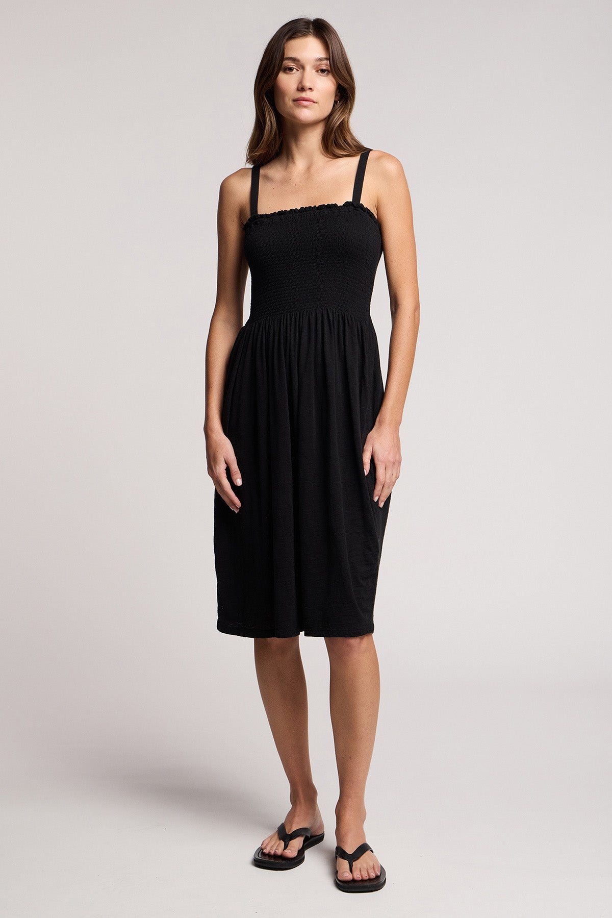 A woman faces forward in the CRESSIDA DRESS by Velvet by Graham & Spencer, a black cotton midi with thin straps ideal for summer, styled with black flip-flops against a plain background.-41371869872321