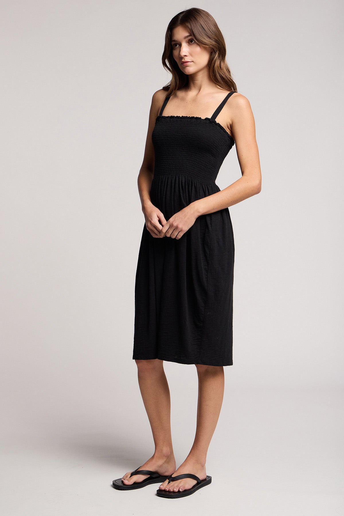A woman stands against a plain background wearing the CRESSIDA DRESS by Velvet by Graham & Spencer, a black sleeveless cotton midi dress with thin straps, paired with black flip-flops.-41371869806785