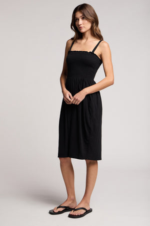 A woman stands against a plain background wearing the CRESSIDA DRESS by Velvet by Graham & Spencer, a black sleeveless cotton midi dress with thin straps, paired with black flip-flops.