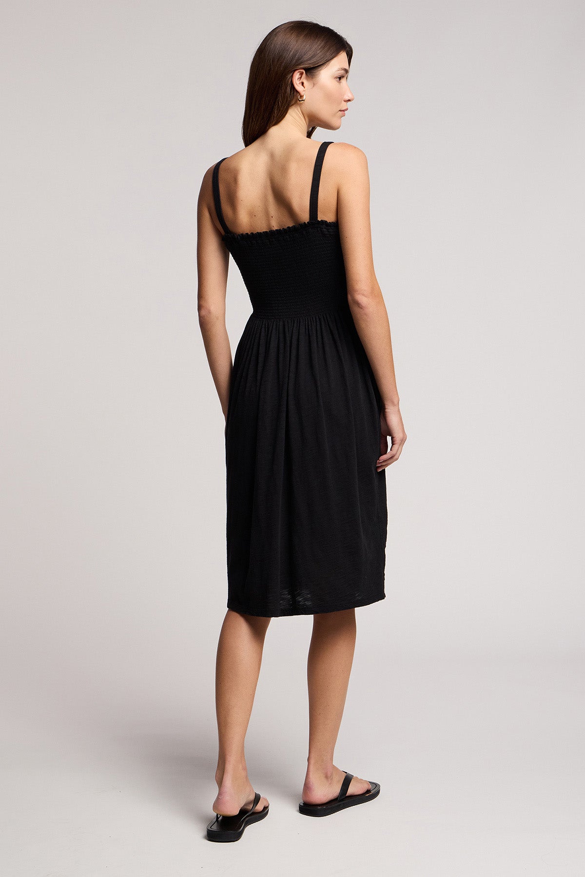 A woman faces away from the camera in a studio, wearing the Velvet by Graham & Spencer CRESSIDA DRESS—a sleeveless black summer dress—paired with black sandals, arms resting at her sides.-41371869708481