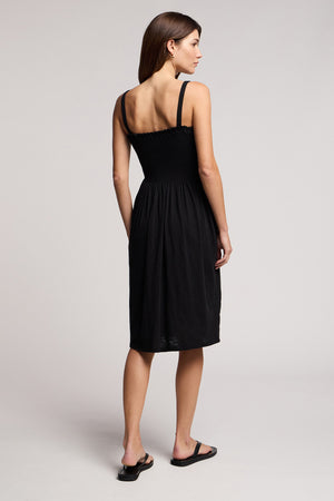 A woman faces away from the camera in a studio, wearing the Velvet by Graham & Spencer CRESSIDA DRESS—a sleeveless black summer dress—paired with black sandals, arms resting at her sides.