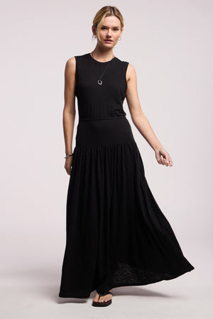 A woman wears the LUNETTA DRESS by Velvet by Graham & Spencer—a sleeveless cotton slub maxi with a fitted top and flared skirt—paired with a necklace and sandals, posed against a plain background.