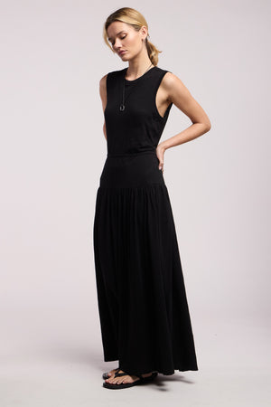 A woman stands against a light background wearing the LUNETTA DRESS by Velvet by Graham & Spencer—a sleeveless black cotton slub maxi—paired with black sandals and a pendant necklace.
