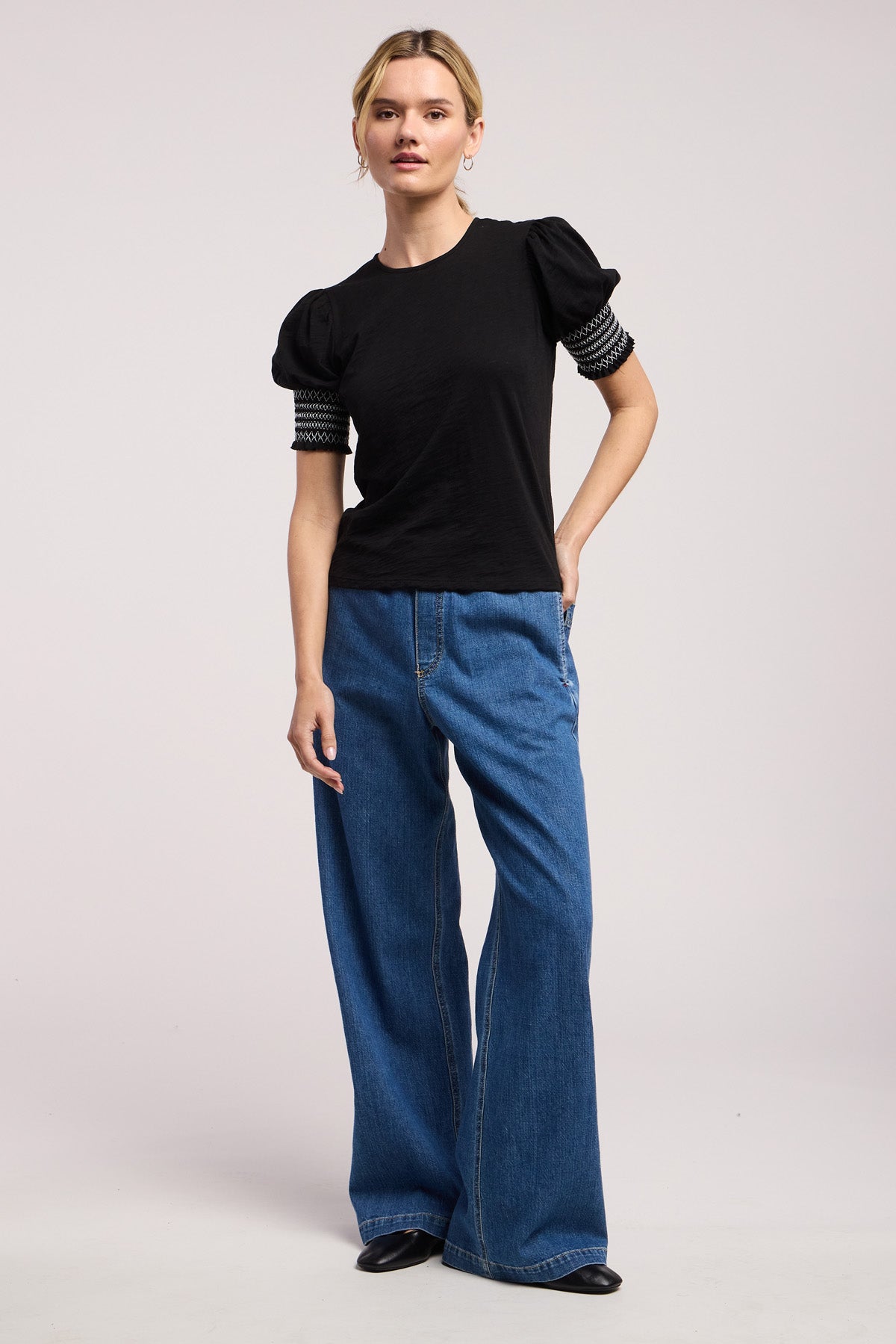 A woman stands against a plain background wearing the SUE TOP by Velvet by Graham & Spencer—a black cotton slub puff-sleeve top—paired with wide-leg blue jeans and black shoes.-41372220948673