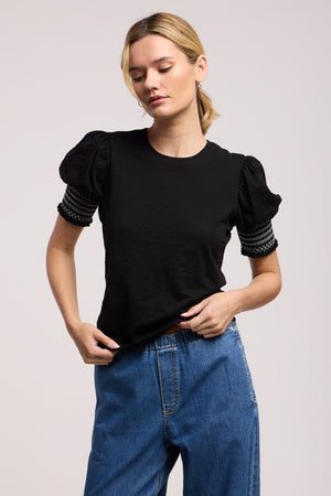 A person wears the SUE TOP by Velvet by Graham & Spencer—a black cotton slub puff-sleeve top with smocked sleeve detailing—paired with blue jeans, standing against a plain light background.
