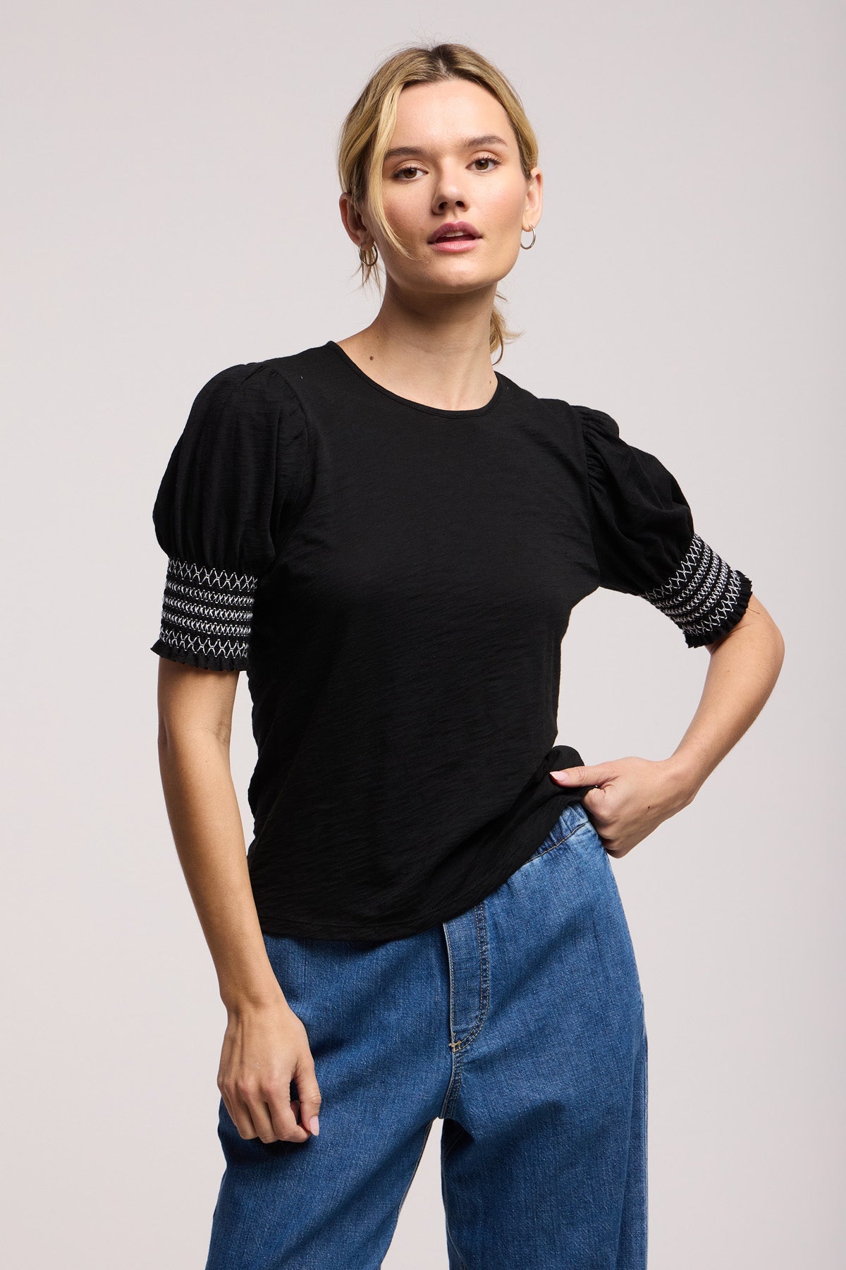 A person wears the SUE TOP by Velvet by Graham & Spencer—a black cotton slub top with patterned sleeve details—paired with blue jeans, standing against a plain light background.