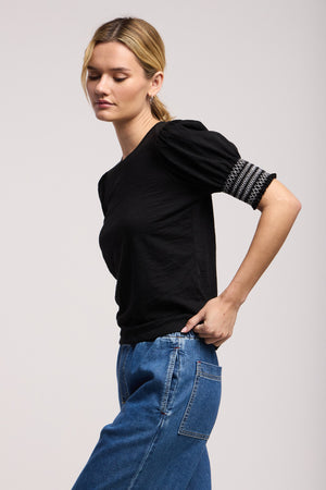 A woman stands sideways against a neutral background, eyes closed, wearing Velvet by Graham & Spencer's SUE TOP—a black cotton slub puff-sleeve style with embroidered detail on one sleeve—paired with blue jeans.