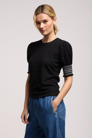 A woman stands against a plain background wearing the SUE TOP by Velvet by Graham & Spencer, a black embroidered top with patterned puff sleeves, paired with blue jeans and one hand in her pocket.