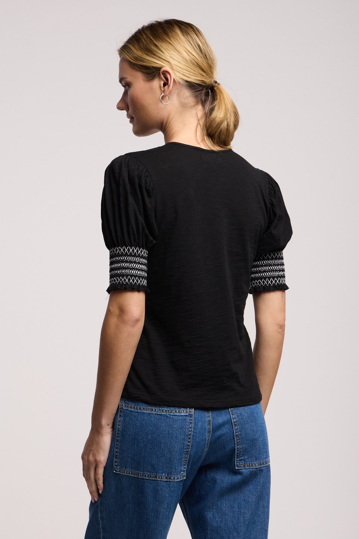 Woman standing with her back to the camera, wearing the SUE TOP by Velvet by Graham & Spencer—a black cotton slub top with patterned trim on puff sleeves—paired with blue jeans.
