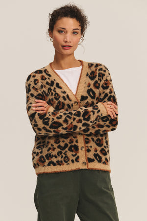 A person stands with arms crossed against a neutral background, wearing the BETSY CARDIGAN by Velvet by Graham & Spencer—a cozy feather yarn leopard print cardigan—layered over a white shirt and olive green pants.