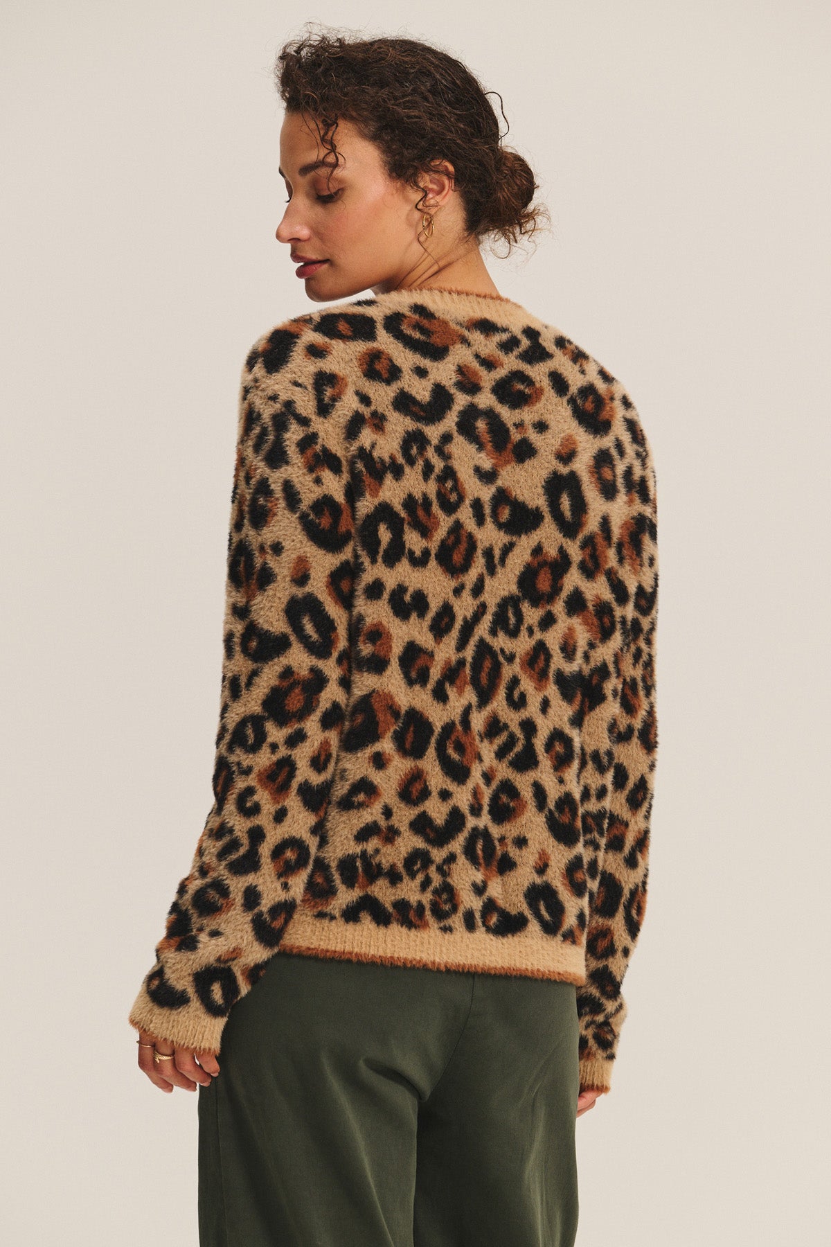 A person stands with their back to the camera, wearing the BETSY CARDIGAN by Velvet by Graham & Spencer in a cozy leopard feather yarn and dark green pants against a plain background.
