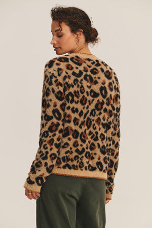 A person stands with their back to the camera, wearing the BETSY CARDIGAN by Velvet by Graham & Spencer in a cozy leopard feather yarn and dark green pants against a plain background.