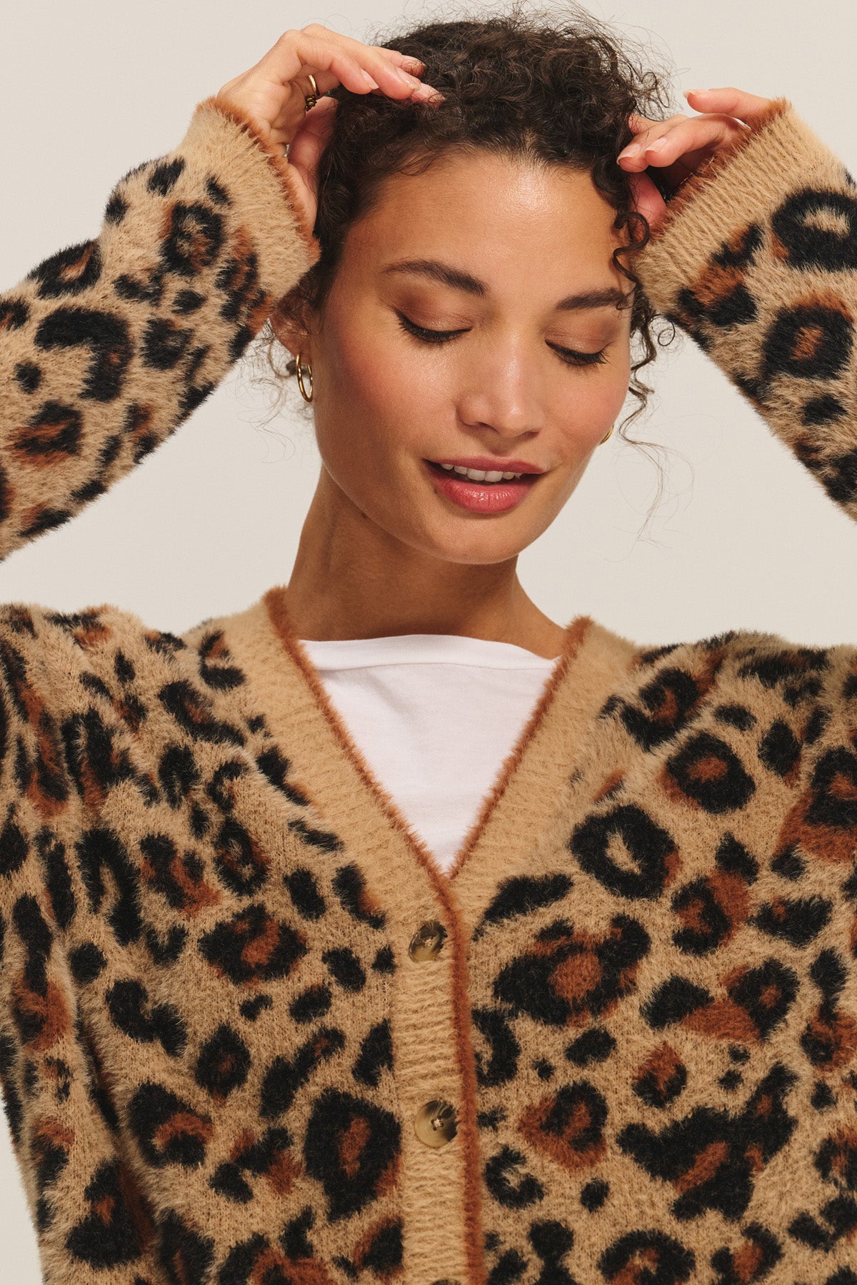 A woman wears the BETSY CARDIGAN by Velvet by Graham & Spencer—a cozy feather yarn piece in tan and black leopard print—over a white top, with her arms raised and eyes closed.