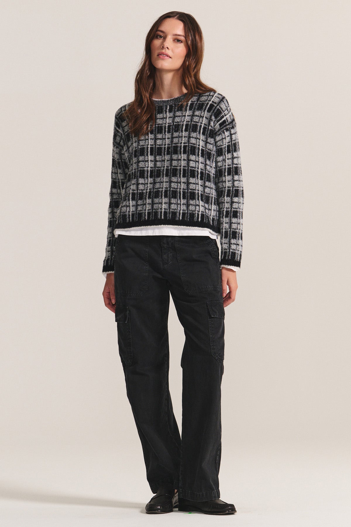 A woman faces forward, wearing a black and white plaid sweater, Velvet by Graham & Spencer MAKAYLA PANT cargo pants with spacious pockets, and black shoes against a plain light background.-40248160780481