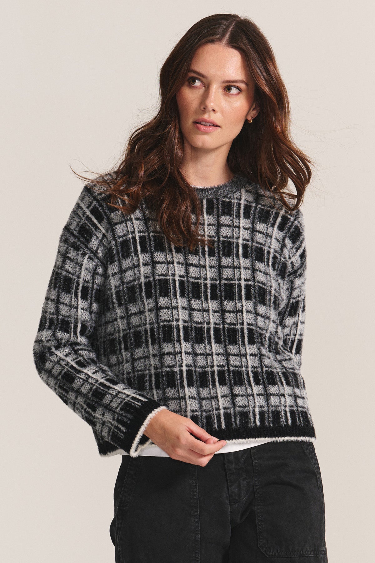 A woman with long brown hair wears the LACEY SWEATER by Velvet by Graham & Spencer— a black and white plaid crewneck—paired with black jeans, standing against a plain light background.-40248075518145