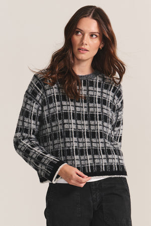 A woman with long brown hair wears the LACEY SWEATER by Velvet by Graham & Spencer— a black and white plaid crewneck—paired with black jeans, standing against a plain light background.