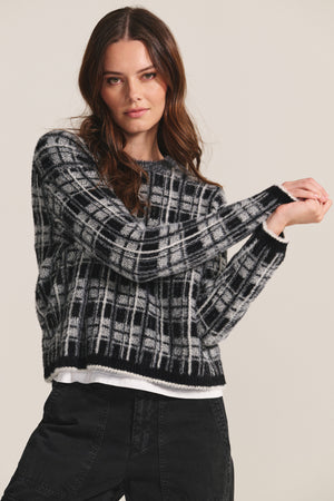 A woman stands against a plain background, arms slightly raised, wearing the LACEY SWEATER by Velvet by Graham & Spencer in black and white jacquard with black jeans.