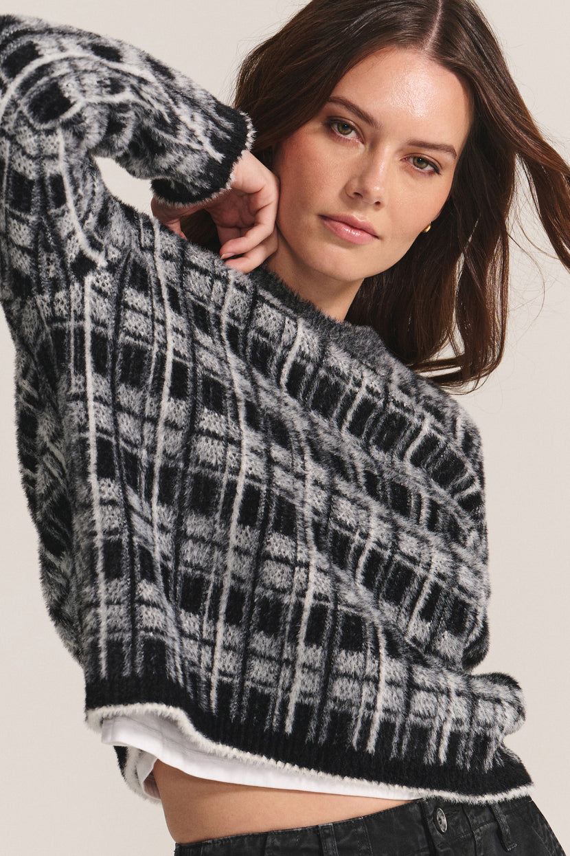 A woman models the LACEY SWEATER by Velvet by Graham & Spencer—a black and gray plaid crewneck—layered over a white shirt with black pants, posing against a neutral background.