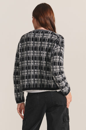 A person with long brown hair is shown from the back wearing the LACEY SWEATER by Velvet by Graham & Spencer, a black and white plaid jacquard crewneck, paired with black pants against a plain background.