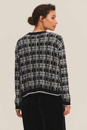A woman with curly hair in a bun is seen from the back wearing the Velvet by Graham & Spencer WANDA CARDIGAN, a black and white plaid sweater, against a plain light background.