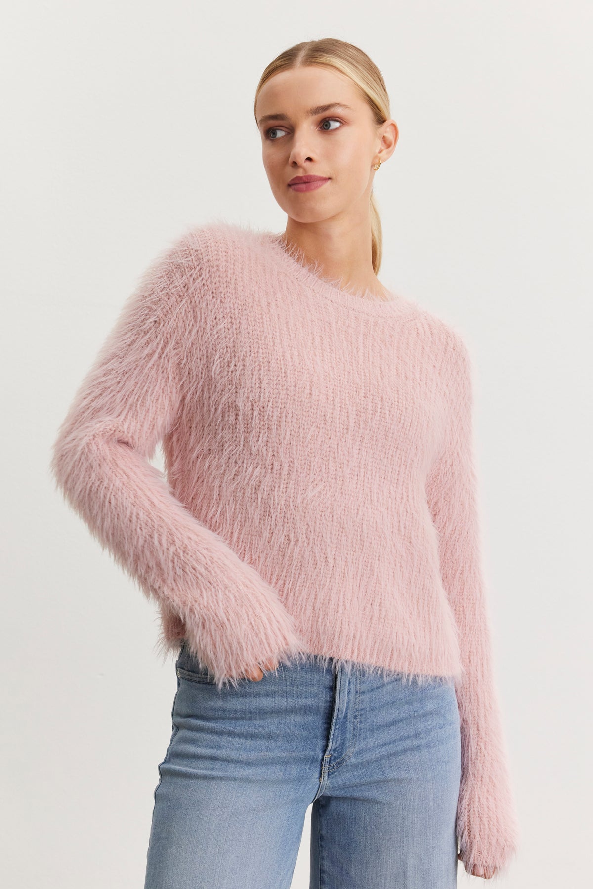   A person wearing the MAYLIE FEATHER YARN SWEATER in light pink by Velvet by Graham & Spencer, paired with light blue jeans, looking slightly to the side against a plain background. 