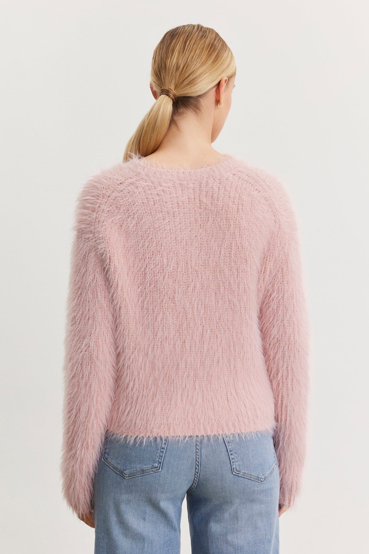   A person with blonde hair in a low ponytail is wearing the MAYLIE FEATHER YARN SWEATER by Velvet by Graham & Spencer, which features a fuzzy pink texture and crew neckline, along with blue jeans, while facing away from the camera. 