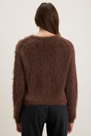 A person is seen from behind wearing the MAYLIE FEATHER YARN SWEATER from Velvet by Graham & Spencer, characterized by its brown feather yarn material and crew neckline, paired with dark jeans against a light background.