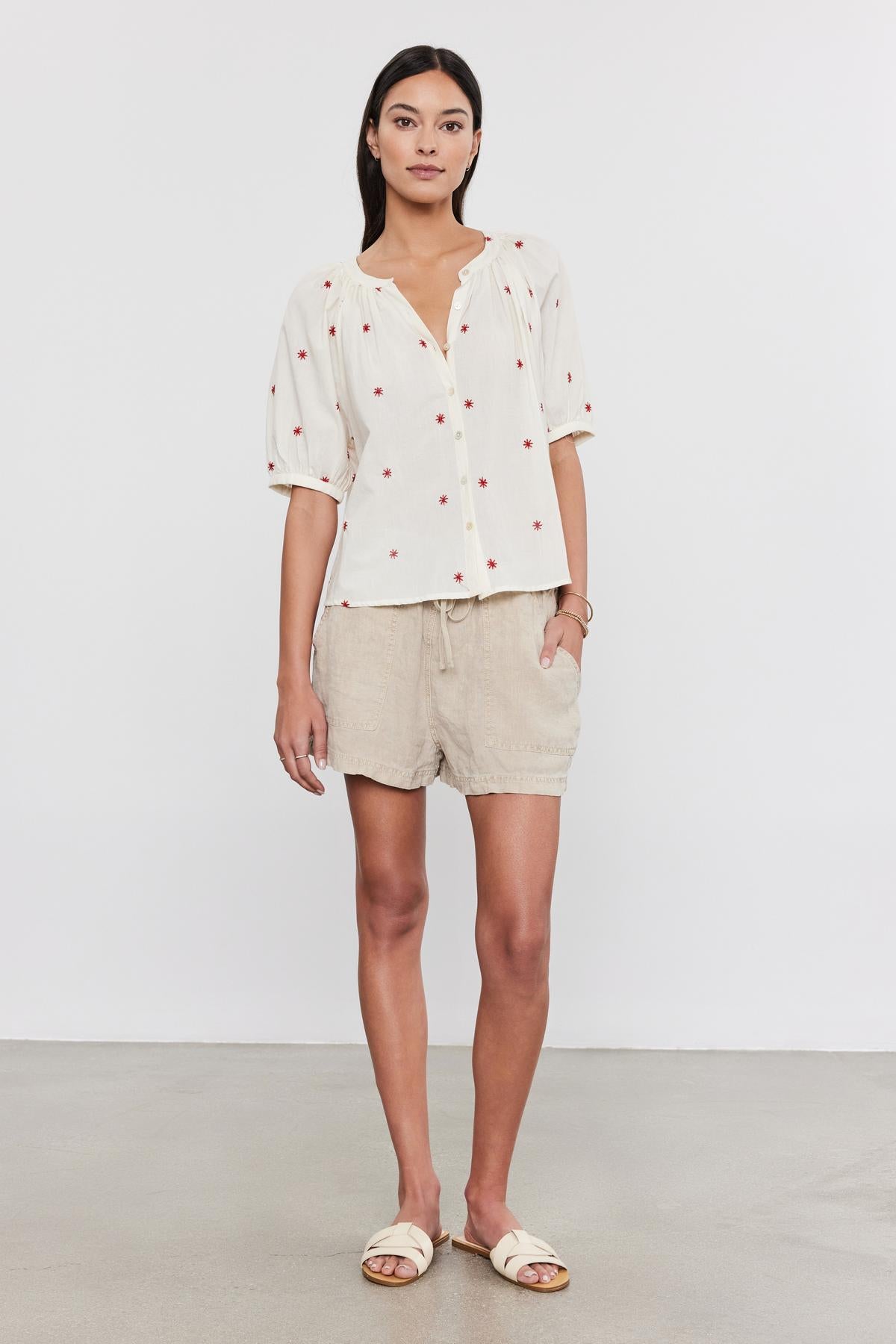 A woman standing in a studio, wearing a white polka-dot Amira top with floral embroidery, beige shorts, and white sandals by Velvet by Graham & Spencer.-36910000275649