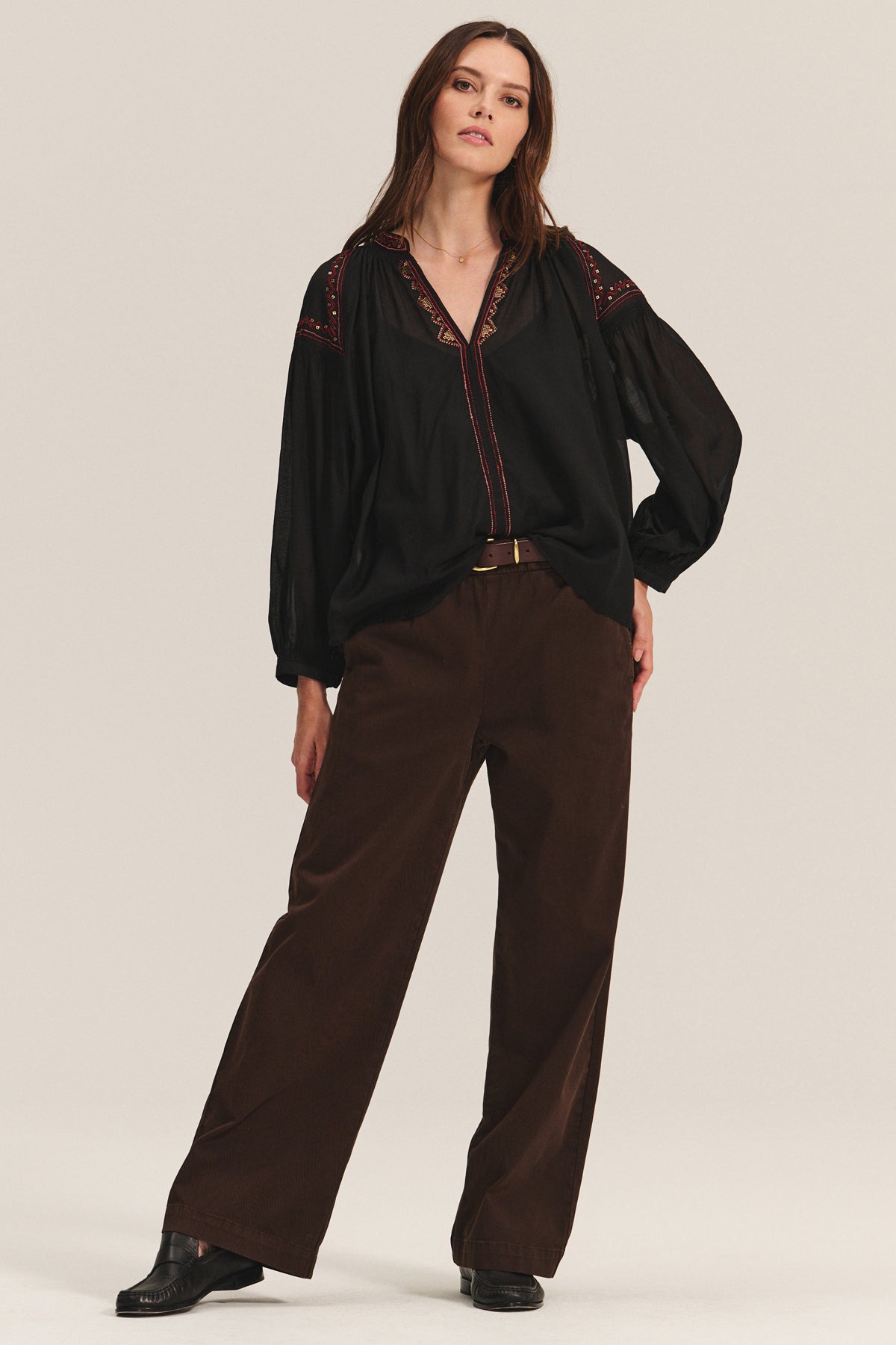 A woman stands against a plain background wearing the BRIDGET TOP by Velvet by Graham & Spencer— a black cotton voile top with red accents—along with brown wide-leg pants and black shoes.-40235004166337