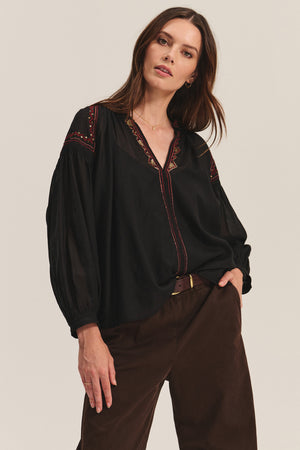A woman wears the BRIDGET TOP by Velvet by Graham & Spencer—a black cotton voile top with beaded embroidered details—paired with brown pants, looking at the camera against a neutral background.