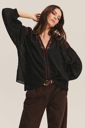A woman models the BRIDGET TOP by Velvet by Graham & Spencer—a loose black cotton voile top with beaded embroidery—paired with brown pants, posing with one hand in her hair against a plain background.