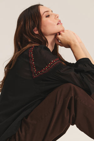 A woman sits with her knees up, resting her chin on her hands. She wears the BRIDGET TOP by Velvet by Graham & Spencer, a black cotton voile top with red and yellow beaded embroidery on the shoulder, paired with brown pants.