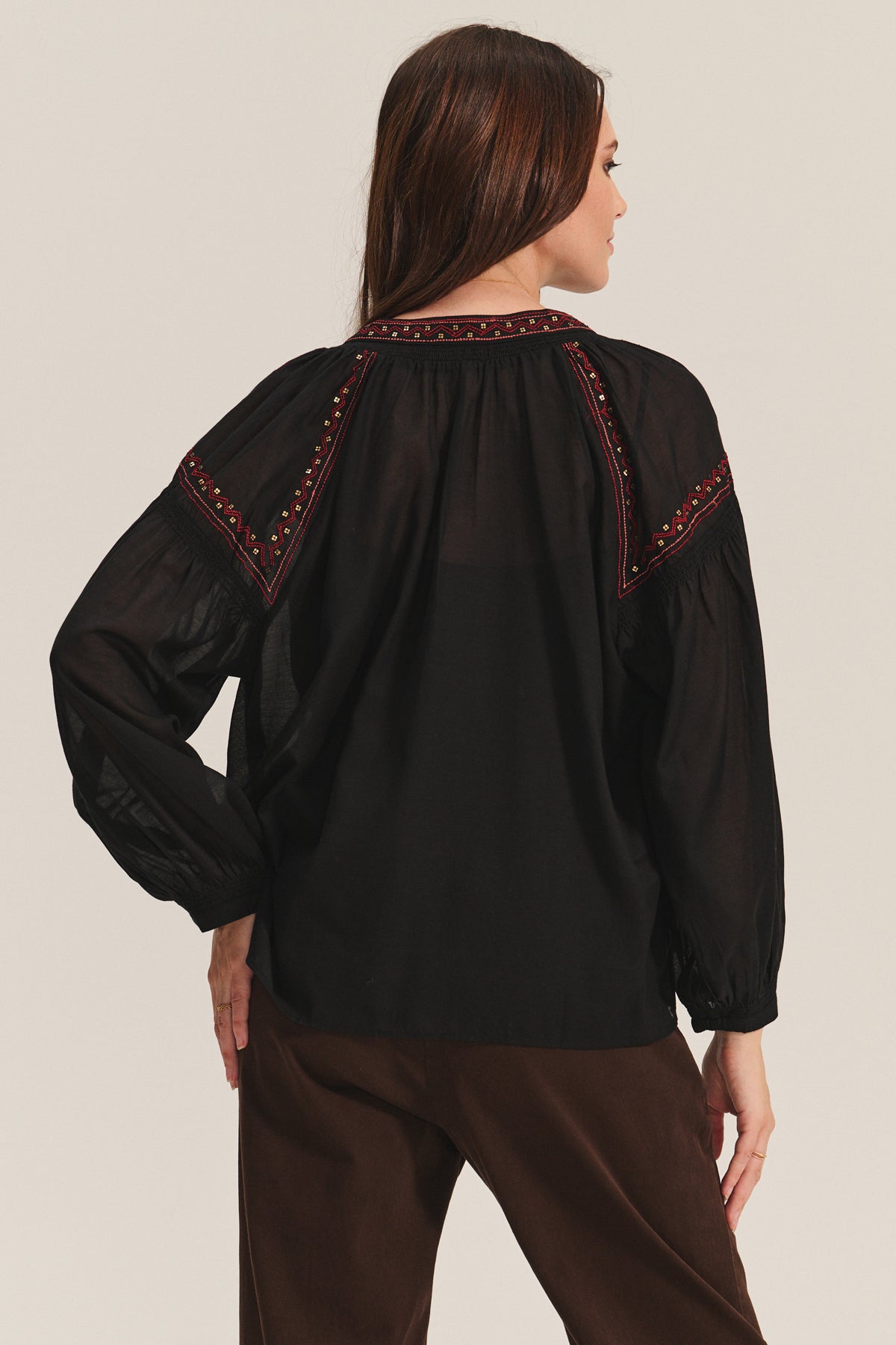 A woman faces away, wearing the BRIDGET TOP by Velvet by Graham & Spencer—a loose-fitting black cotton voile top with beaded embroidery on the shoulders and yoke—paired with brown pants.
