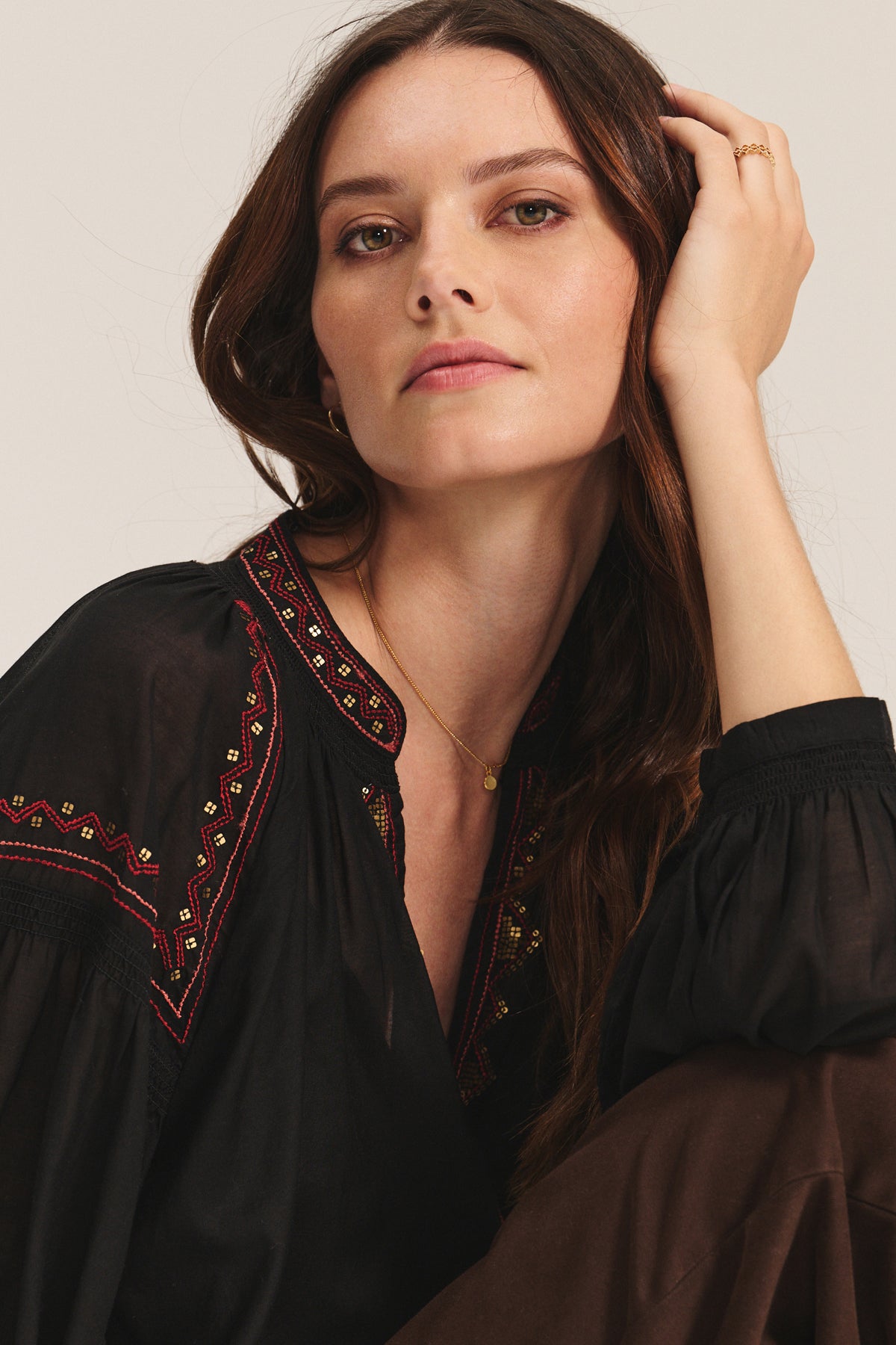 A woman with long brown hair wears the BRIDGET TOP by Velvet by Graham & Spencer— a black cotton voile top featuring red, gold, and beaded embroidered details—resting her head on her hand and looking at the camera against a plain background.-40235004002497