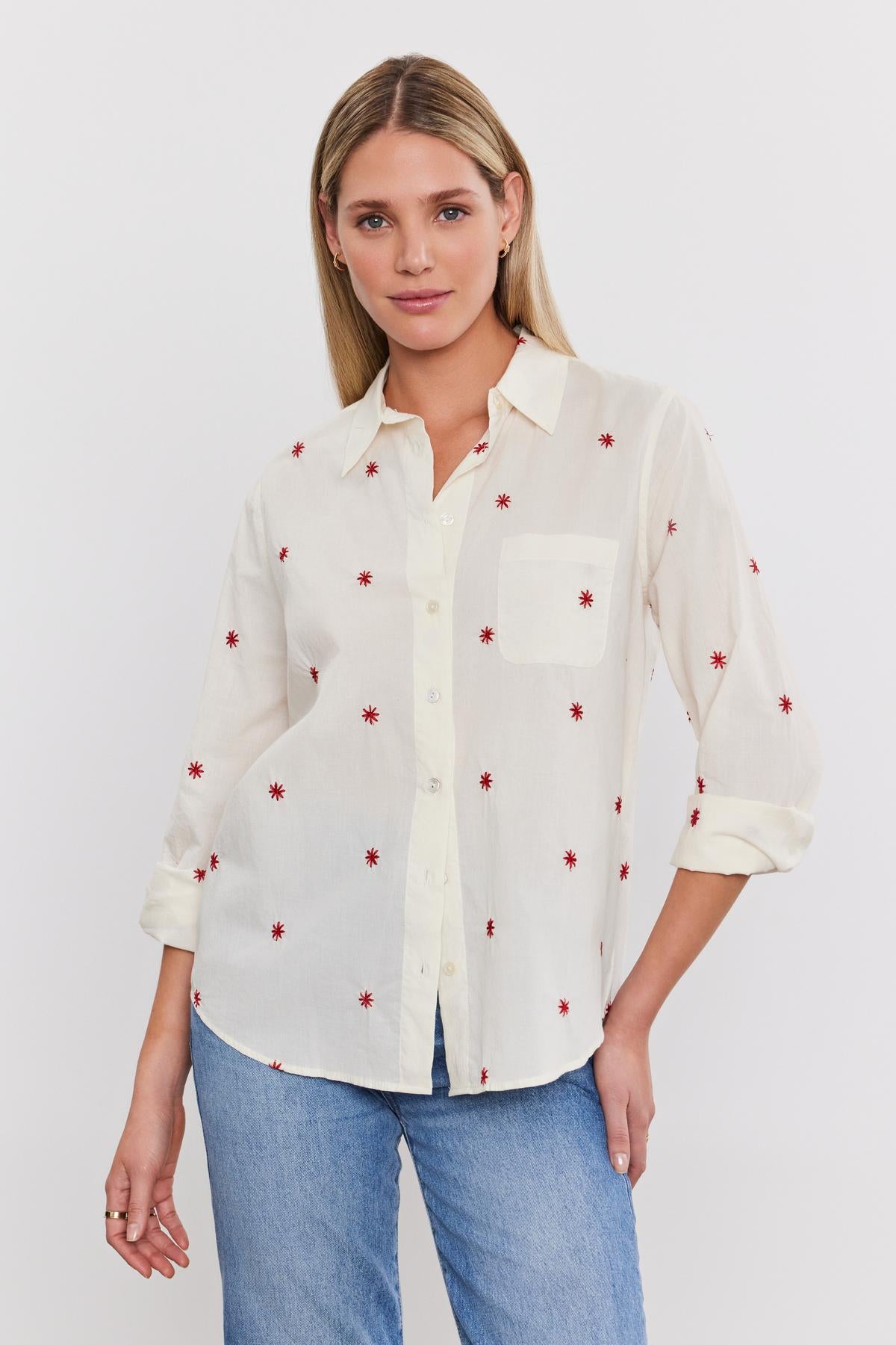   A woman with long blonde hair, wearing the EMBERLY SHIRT by Velvet by Graham & Spencer—a white button-down shirt with red star patterns—and blue jeans, stands against a white background. 